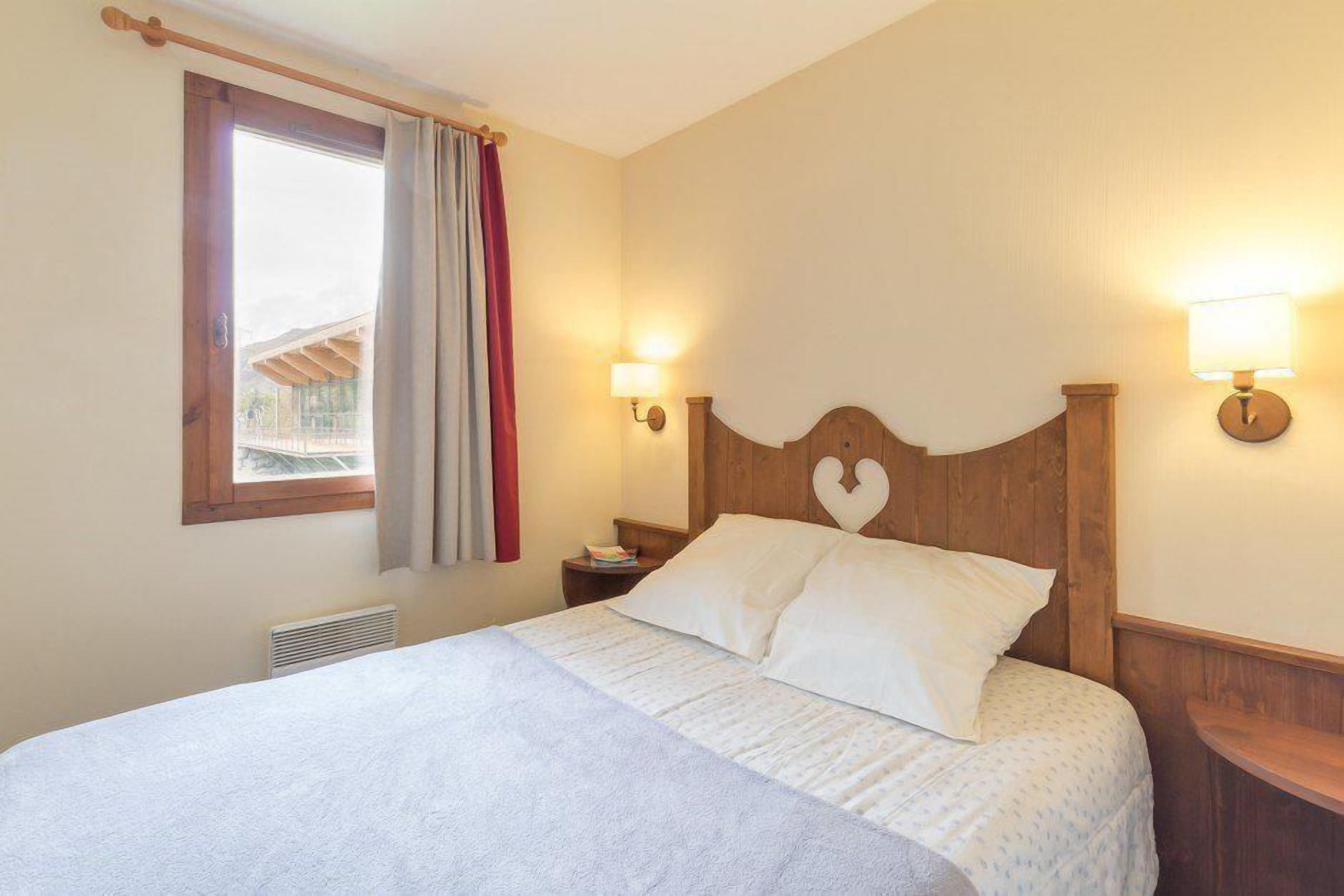 A bedroom with a wooden headboard, two bedside lamps, a window with curtains, and a bed with white bedding.