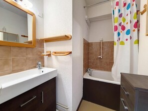 Ein Badezimmer mit einem weißen Waschbecken, einem Spiegel, einem Holzregal, einer Dusche mit bunter Duschvorhang und einem dunklen Schrank.