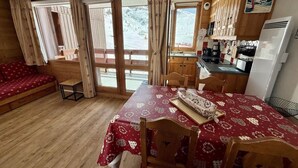 Een gezellige kamer met een houten vloer, een eettafel met een rode tafelkleed, een bank en een keukenhoek met een koelkast en kastjes.