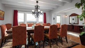 Een eetkamer met een grote tafel en gestreept beklede stoelen, een kroonluchter en uitzicht door de ramen.