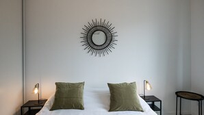 Un dormitorio con una cama, dos almohadas verdes, dos mesitas de noche con lámparas y un espejo circular montado en la pared.