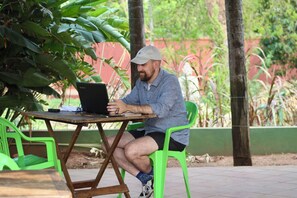 Una persona sentada en una mesa de madera con una computadora portátil, usando una gorra y pantalones cortos, en un entorno exterior con sillas verdes y plantas.