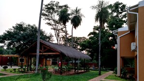 A rustic outdoor seating area with a thatched roof, surrounded by lush greenery and palm trees. - Your Personal Paradise in ZenGarden: Cozy Vacation Home for Couples in Paraguay (Ñemby)