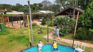 A swimming pool with a ladder, surrounded by a grassy yard and a wooden structure. - Your Personal Paradise in ZenGarden: Cozy Vacation Home for Couples in Paraguay (Ñemby)