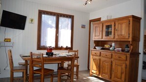 Een eetgedeelte met een houten tafel en stoelen, een houten kast met glazen deuren, een aan de muur bevestigde televisie en een raam met gordijnen.