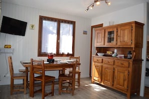 Ein Essbereich mit Holztisch und Stühlen, einer Holzvitrine mit Glastüren, einem an der Wand montierten Fernseher und einem Fenster mit Vorhängen.