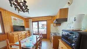 Ein Holzinnenraum mit Esstisch, Stühlen, einem Fernseher, einer Küchenzeile mit Mikrowelle und Toaster sowie Blick durch ein Fenster.