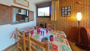 Ein Essbereich mit Holztisch, Stühlen und einer Tischdecke. Es gibt ein Fenster mit Vorhängen, einen an der Wand montierten Fernseher und eine Lampe.