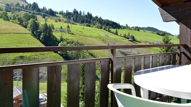 Un balcon doté d’une balustrade en bois, une table blanche et une vue sur une vallée verdoyante, avec des arbres et une montagne en arrière-plan.