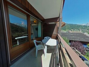 Un balcón con mesa y sillas, vista a un paisaje montañoso y fachada de un edificio de madera.