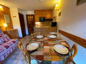 A cozy dining area with a round table set for four, wooden chairs, and a kitchen in the background. - 300m from resort center, 1st floor, balcony, tv, 23m², Le Grand Bornand (Le Grand-Bornand)