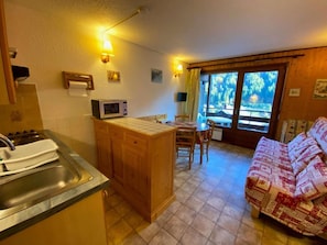 A compact kitchen with a sink, microwave, and wooden cabinets. There is a dining area with a table and chairs, and a sofa with a patterned cover. - 1st floor, balcony, tv, 23m², Le Grand Bornand (Le Grand-Bornand)