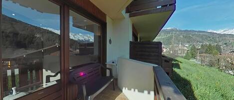 A balcony with a view of snow-capped mountains, a wooden deck, and a small table with a chair.
