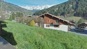A modern chalet with a sloped roof, wooden exterior, and a balcony. It is surrounded by lush green fields and mountains in the background.