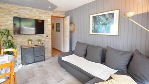 A modern living room with a grey sofa, a flat-screen TV mounted on a stone wall, a dining area with a wooden table and chairs, and a painting on the wall.