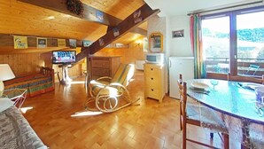 Una habitación acogedora con pisos de madera, una mecedora, una mesa de comedor con sillas, un televisor y una ventana con cortinas.