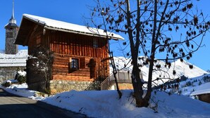 Une cabane en bois, dont le toit est recouvert de neige, avec une cheminée en pierre, et un ciel bleu clair en arrière-plan.