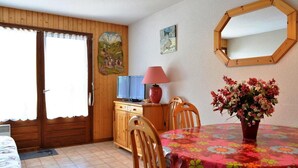 Une pièce avec une porte en bois, un téléviseur posé sur un meuble en bois, une table recouverte d’une nappe à motifs floraux et un miroir accroché au mur.
