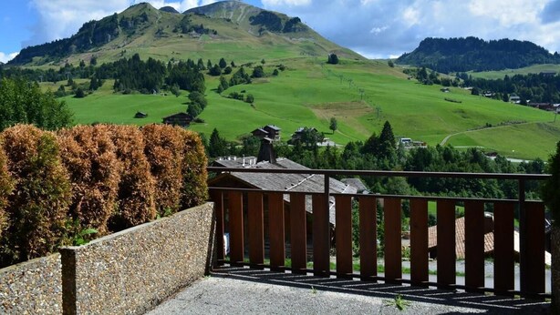 Une vue pittoresque depuis un balcon doté d’une balustrade en bois, donnant sur une vallée verdoyante, avec des collines et une montagne en arrière-plan.