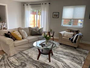 Un salon avec un canapé beige, une table basse en verre et un fauteuil beige. Une fenêtre offre une vue sur un mur de briques et un jardin.