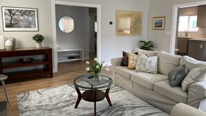 Una sala de estar moderna con un sofá color beige, una mesa de centro de vidrio y una alfombra con diseño de mármol. Se observa una consola de madera con objetos decorativos, un cuadro en la pared y un espejo redondo.