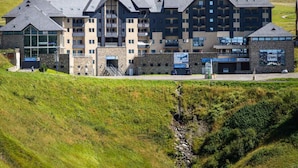 A multi-story hotel with a modern architectural design, featuring a mix of stone and glass facades, located in a hilly area with a view of the surrounding landscape. - 100m from ski slopes, 1st floor, jacuzzi, hammam, parking, tv, ski locker, 41m², Peyragudes (Germ)