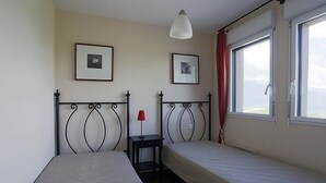 Une chambre avec deux lits simples, une petite table surmontée d’une lampe rouge et deux tableaux encadrés accrochés au mur.