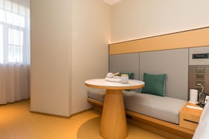 A modern hotel room with a round wooden table, a sofa with a green pillow, and a bedside table with a lamp.