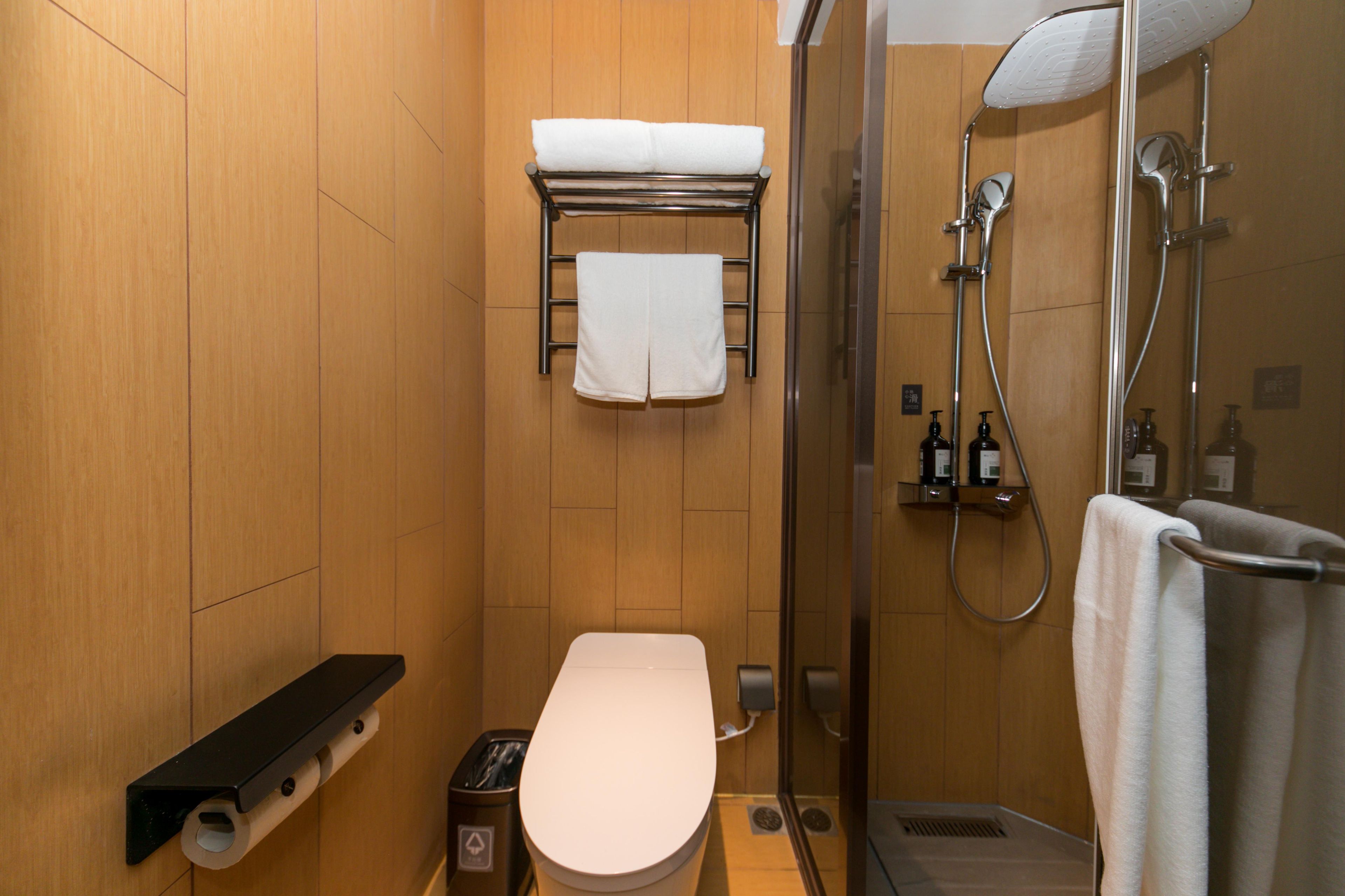 A compact bathroom with a toilet, a shelf, a towel rack, and a shower area.