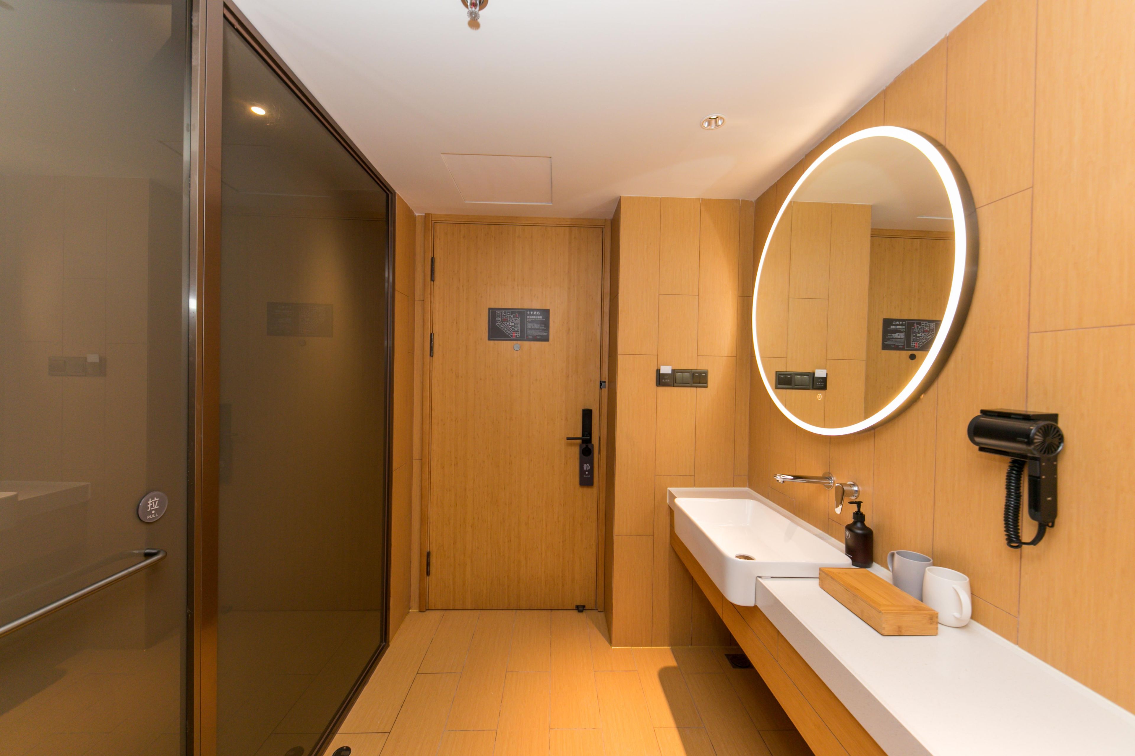 Une salle de bain moderne avec une porte en verre, un meuble-lavabo en bois, un miroir avec éclairage intégré et un sèche-cheveux.