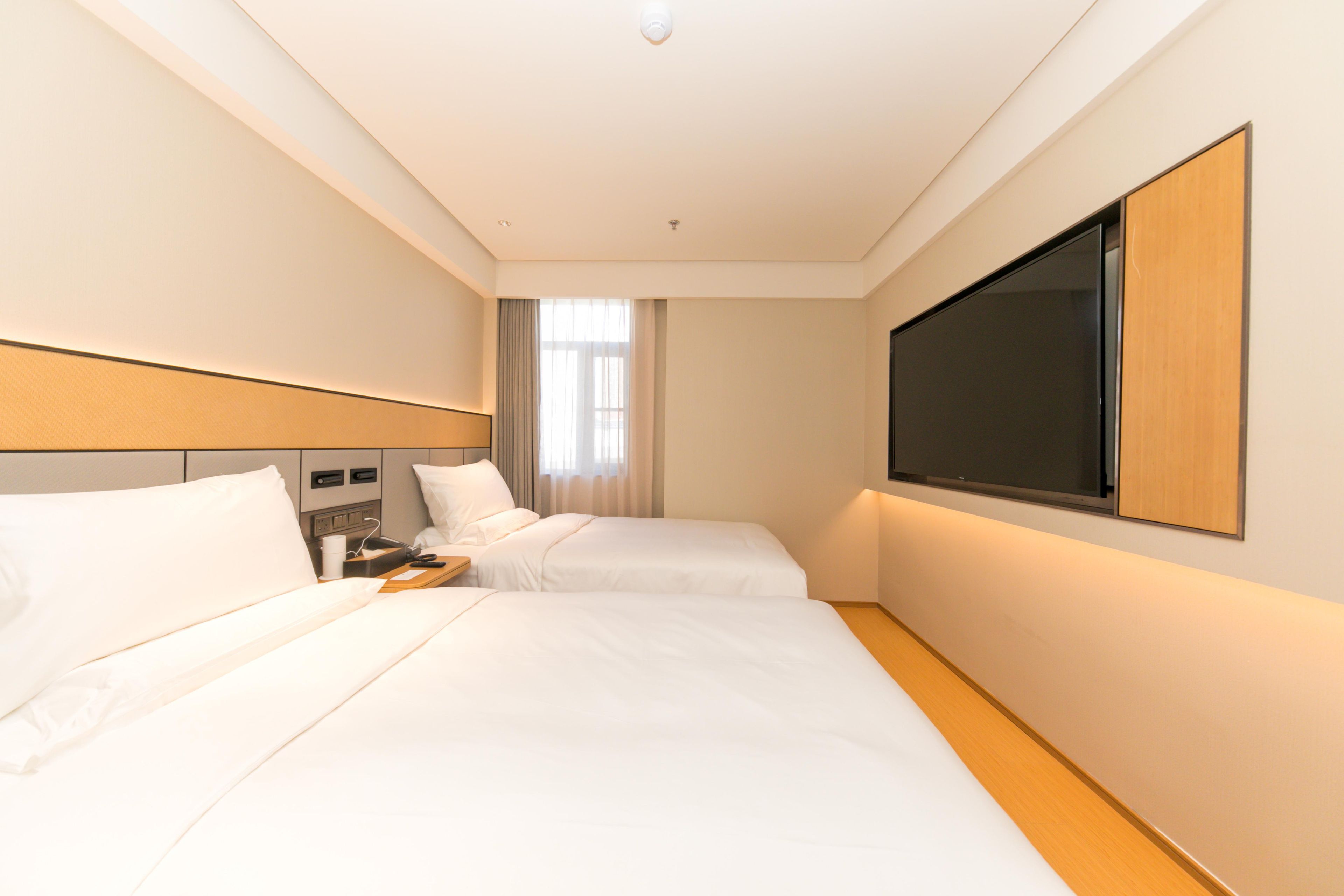 A hotel room with two beds, a flat-screen TV, a bedside table, and a window with curtains.
