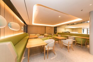 Un interior moderno con mesas de madera y asientos verdes, una zona de cocina y un reloj visible en la pared.
