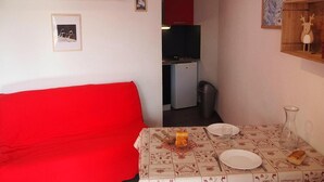 Ett kompakt vardagsrum med en röd soffa, ett matbord med ett mönstrat bordsduks och en köksdel med ett kylskåp och en liten spis.