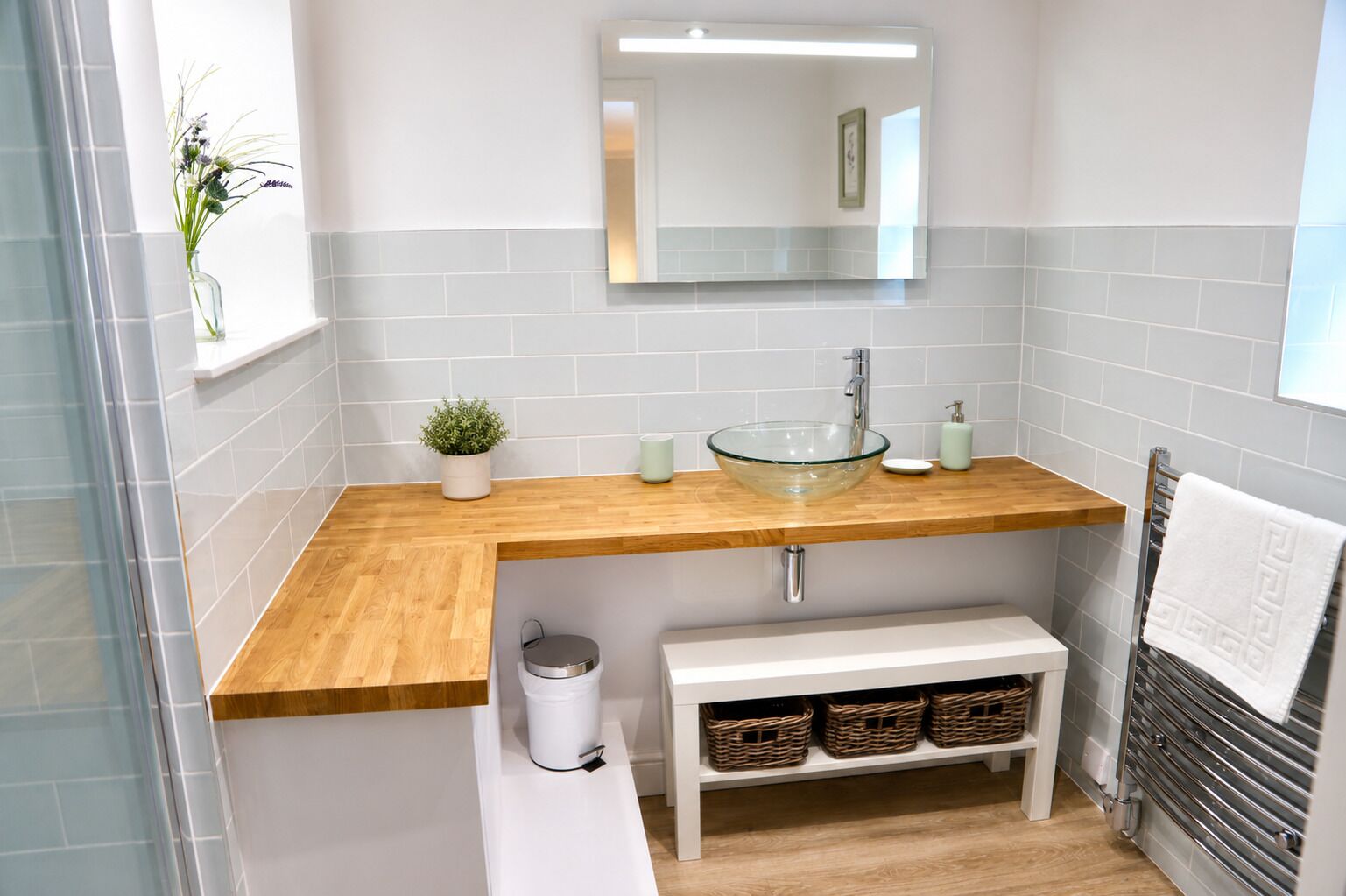 A bathroom with a glass bowl sink, wooden countertop, white tiled walls, a mirror, a small potted plant, a trash bin, and a towel rack.