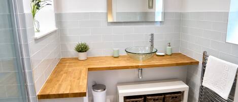 A bathroom with a glass bowl sink, wooden countertop, white tiled walls, a mirror, a small potted plant, a trash bin, and a towel rack.