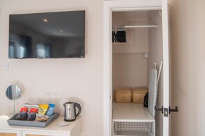 A hotel room with a flat-screen TV, a small mirror, a tray with snacks and a kettle, and a neatly organized closet with hanging clothes and a folded towel. - Bridgeside Rooms (Telford)