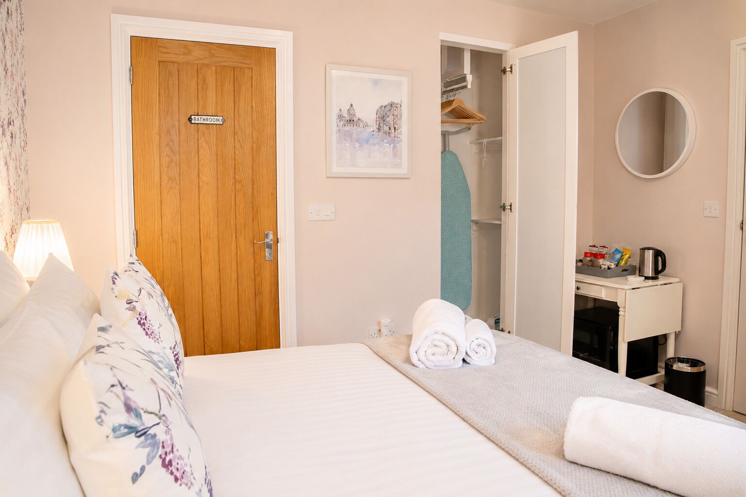 A hotel room with a bed, a bathroom sign, a towel rack, a mirror, and a small table with a kettle.