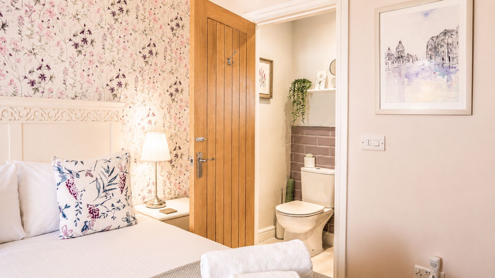 A bedroom with a bed, pillows, a bedside table with a lamp, a wooden door, and a bathroom with a toilet and shower.
