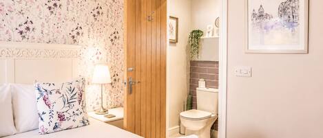 A bedroom with a bed, pillows, a bedside table with a lamp, a wooden door, and a bathroom with a toilet and shower.