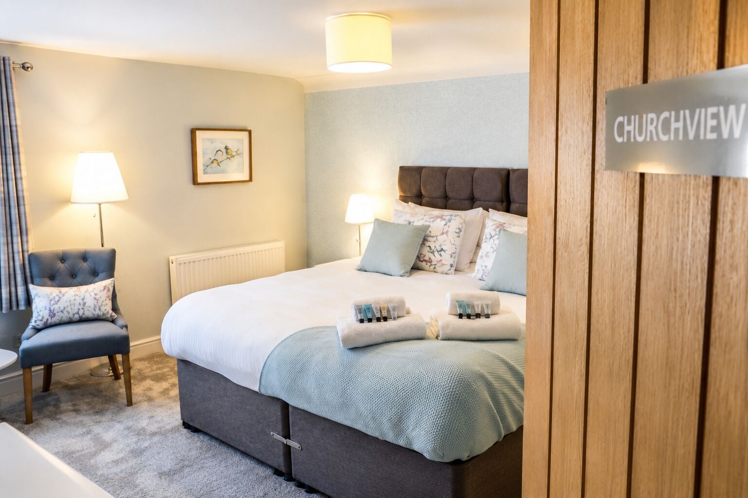 A bedroom with a bed, two bedside lamps, a chair, a radiator, and a sign reading 'CHURCHVIEW'.