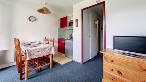 Una stanza compatta con un tavolo da pranzo, sedie, una TV su un comò, angolo cottura con mobili rossi e una porta che dà su un'altra stanza.