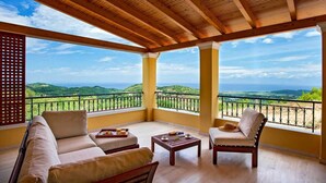 Un balcon doté d’un plafond en bois, de canapés beiges, d’une table basse en bois et offrant une vue sur des collines verdoyantes et la mer.