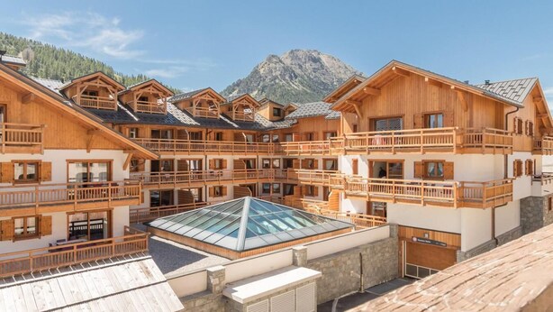 A multi-story wooden chalet with balconies, a glass-roofed terrace, and a mountainous backdrop.