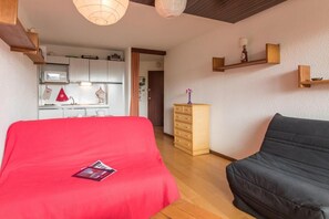 A compact bedroom with a red bed, a black sofa, a wooden dresser, and a kitchenette in the background. - 220m from resort center, 5th floor, view village, balcony, parking, tv, 25m², Montgenèvre (Montgenèvre)