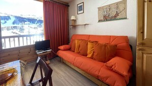 Une pièce chaleureuse avec un plancher en bois, un canapé orange vif, une petite table avec un ordinateur portable, une fenêtre avec des rideaux rouges et une carte encadrée accrochée au mur.