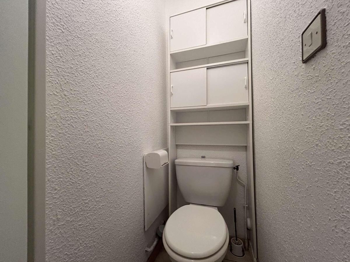 A small, white bathroom with a toilet, a shelf above the toilet, and a wall-mounted light switch.
