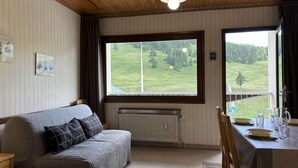 Ein gemütliches Zimmer mit Holzbalkendecke, einem großen Fenster mit Blick auf eine grüne Landschaft, einer Couch mit Kissen, einem Heizkörper und einem Esstisch mit Tellern und Gläsern.