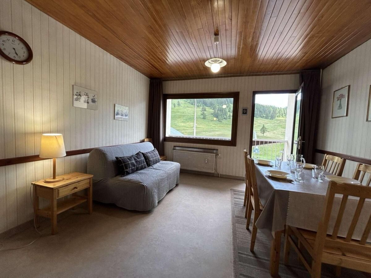 A cozy room with a wooden ceiling, a sofa, a dining table set for four, and a window offering a view of a green landscape.