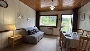 Ein gemütliches Zimmer mit Holzbalkendecke, einer Couch, einem Esstisch für vier Personen und einem Fenster mit Blick auf eine grüne Landschaft.
