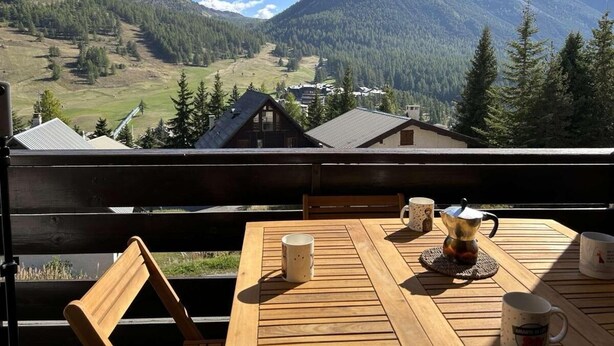 Una mesa de madera con dos tazas y una tetera en un balcón que da a un paisaje montañoso con casas.